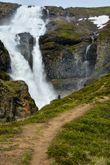 Obraz premium Fresh clean small waterfall in Iceland in summer with loads of water flowing between rocks and unrecognisable person by the waterfall, snow in the background