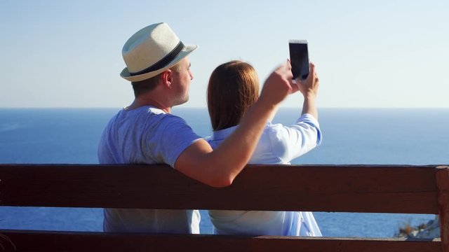 Young couple in love relaxing on bench at edge of cliff doing selfie on mobile phone. Husband and wife on honeymoon enjoying breathtaking view of blue Mediterranean sea making photos on cellphone