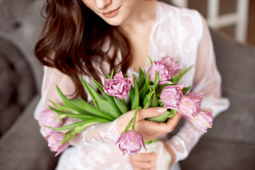 Beautiful young woman in the white dress with flowers tulips in hands on bed at home.