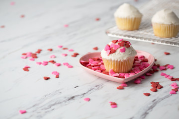 Vanilla Valentine Cupcake with White Frosting and Heart-shaped Sprinkles on Wire Cooling Rack with One Isolated in Front in Pink Heart-Shaped Dish and Heart-Shaped Sprinkles Scattered on Countertop