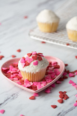Vanilla Valentine Cupcake with White Frosting and Heart-shaped Sprinkles on Wire Cooling Rack with One Isolated in Front in Pink Heart-Shaped Dish and Heart-Shaped Sprinkles Scattered on Countertop