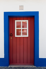 Red wooden door with four small windows. White wall with blue painting around the door. Number plate above the door.
