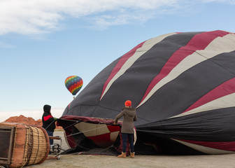 Obraz premium Two women in warm winter clothes prepare a hot air balloon as another balloon lifts into the sky in the distance.