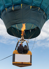 Two people ride in the basket of a hot air balloon as the flame from the burner heats the air inside. A sign on the basket is blank.