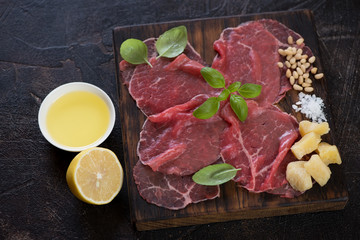 Rustic wooden serving board with carpaccio beef and condiments, studio shot on a dark brown stone surface