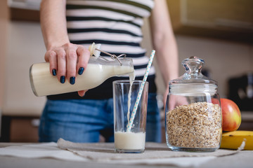Woman pours organic oat milk from a bottle into a glass on a table in the kitchen. Diet healthy...