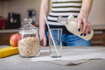 Woman pours organic oat milk from a bottle into a glass in the kitchen. Diet healthy vegetarian...