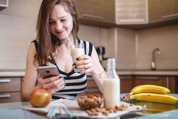 Woman drinking organic almond milk holding a phone in her hand in the kitchen. Diet healthy vegetarian product