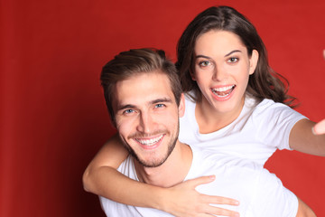Happy young couple while sitting on back of content man isolated over red background, taking a selfie.
