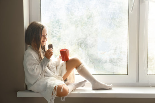 Cute little girl wrapped in warm plaid with cup of hot cocoa drink eating chocolate on windowsill at home