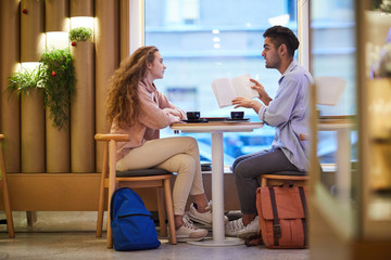 Fototapeta premium Two teenage groupmates or dates sitting in college cafe before lesson and discussing notes in copybook
