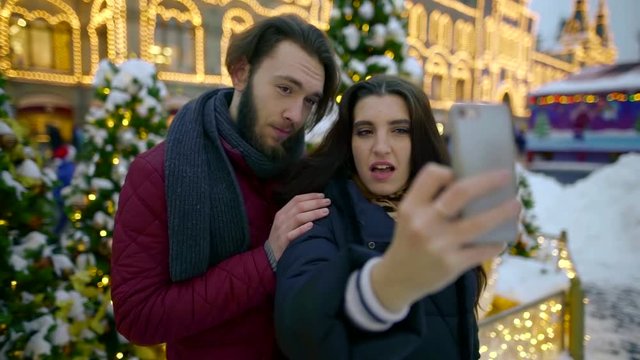 Emotional and happy couple taking couple-selfies on a smartphone against beautiful christmas decorations and trees in a park.