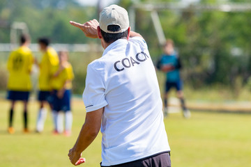 Back of football coach wearing white COACH shirt at an outdoor sport field coaching his team during a game