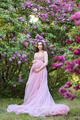 Obraz na płótnie Canvas Beautiful young pregnant woman in a luxurious pink dress walking in a park of blossoming lilacs.