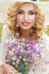 Naklejka premium Curly blonde in a white hat walks in a park with a bouquet of field violet flowers..