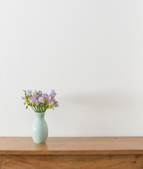 Purple freesia flowers in green vase on wooden table against white wall with copy space (selective focus)