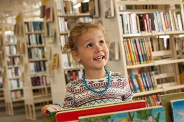 Preschooler little girl smiling and reading book in library. Kid with books near a bookcase. Happy, cheerful and cute girl with german books. Preschool concept. Children creativity