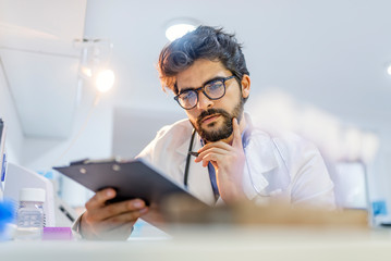Fototapeta premium Doctor, in white uniform, sits at her desk and writes something to her notebook.