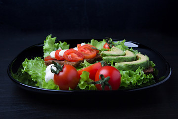 Salad and mozzarella, healthy sandwiches from black unleavened rye bread with fresh tomatoes and mozzarella cheese, with ripe avocados on a glossy black plate.