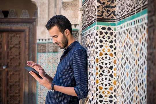 Young Muslim man in casual clothing smiling and working on tablet in traditional Arabian ambient