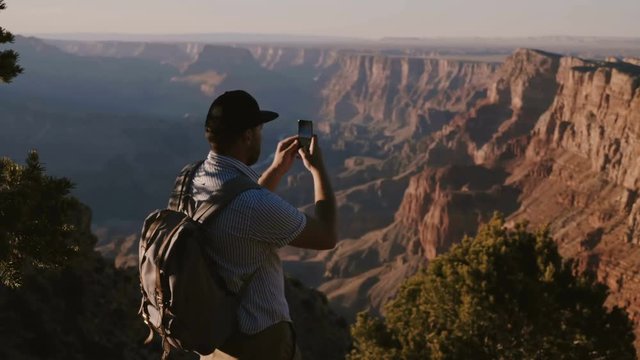 Slow motion happy young tourist man with backpack taking smartphone photo of epic summer sunset over Grand Canyon USA.