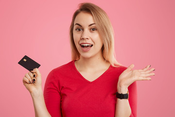 Naklejka premium Headshot of positive hesitant light haired European woman dressed in red jumper, holds credit card, has clueless expression, poses over pink background, wants to make payments or transaction.