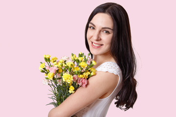 Obraz na płótnie Canvas Close up shot of attractive woman with make up, natural beauty, gentle smile, has long straight hair, carries flowers, recieves bouquet on 8 March, wears stylish dress, isolated over rosy background