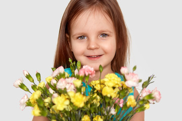 Obraz na płótnie Canvas Good looking little kid with charming smile, holds flowers, has pleased facial expression, enjoys nice day, isolated over white background. Blue eyed small child celebrates birthday. Indoor shot