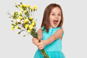 Photo of overjoyed pleased small cute child exclaims happily, dressed in blue dress, holds bouquet of flowers, focused aside, isolated over white background. Childhood and positiveness concept