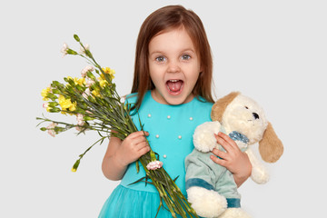 Obraz na płótnie Canvas Image of overjoyed blue eyed small child holds her favourite toy and flowers, happy to recieve present on birthday, opens mouth widely, dressed in festive dress, isolated over white studio wall