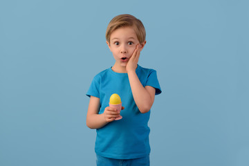 Child and Easter. Smiling blond boy, 6 years old, is holding a yellow egg in his hand. Easter time. Waist up portrait. Blue studio background.