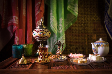 Arabian tea in glass with eastern snacks on vintage wooden surface. Eastern tea concept. Low light lounge interior with carpet. Empty space.