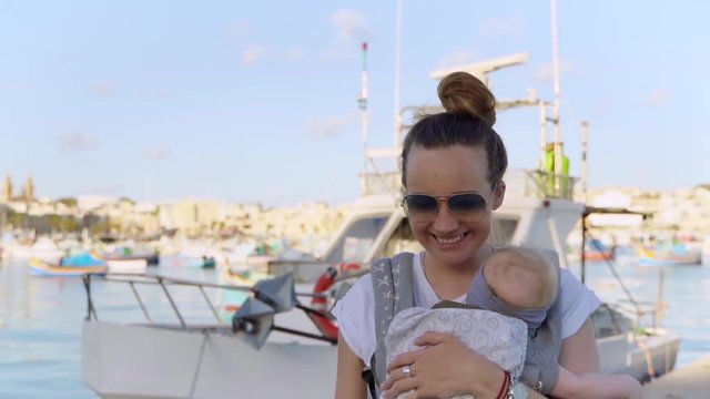 A middle-plan of a young mother with bun hairstyle walking with her little baby in a sling in front of traditional fishing boats Luzzu in Marsaxlokk, Malta