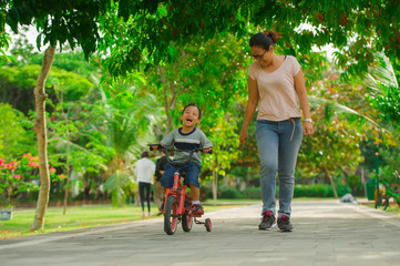 lifestyle portrait of Asian Indonesian mother and young happy son at city park having fun together the kid learning bike riding and the woman running after the child