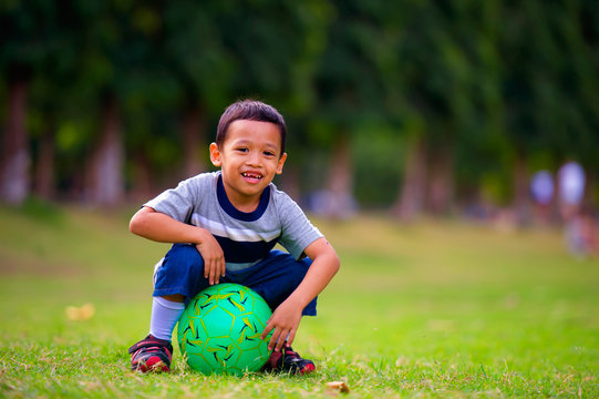 outdoors portrait at grass city park of 5 years old Asian Indonesian child smiling happy and cheerful  sitting on ball laughing playful isolated on green trees background