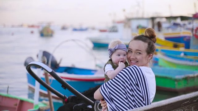 A pretty mother sitting on the bench and talking with her yearling baby near the sea in Marsaxlokk, Malta