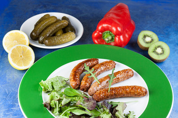 PLATO DE SALCHICHAS DE CARNE CON PIMIENTO ROJO, PEPINILLOS EN VINAGRE, KIWIS Y LIMONES SOBRE FONDO AZUL