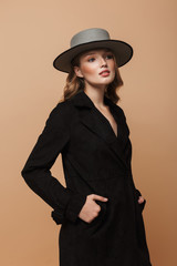 Young gorgeous woman with wavy hair in black coat and gray hat dreamily looking aside standing over beige background