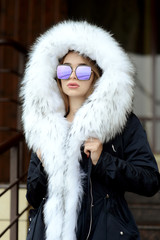 a girl in a black jacket with a white fur collar and glasses posing on the street
