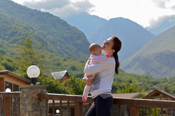 Fototapeta na wymiar Happy mother with a small child standing on the terrace of a country house surrounded by mountains on a summer day.
