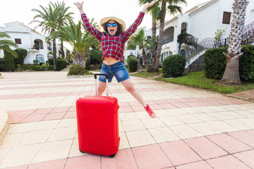 Travel, tourism, emotions and people concept - happy young woman jumped in a hat and sunny glasses near her red suitcase