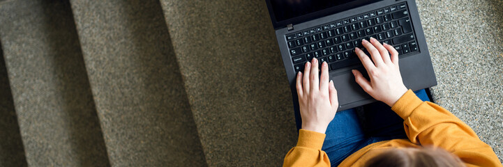 Young female college student sitting on stairs at school, writing essay on her laptop. Education concept. View from above.