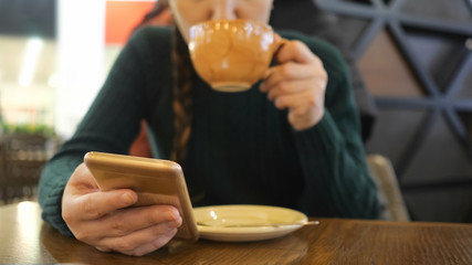 phone in the hand of the girl in focus, woman is drinking coffee on the blurred background.