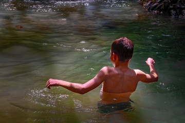 Boy is swimming in a forest river in the shade of tropical trees. Rays of the sun through leaves fall on the body. Rear view.