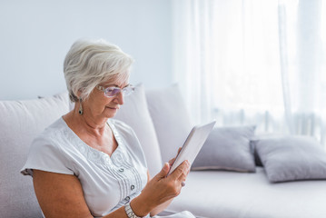 Senior woman reading a message, e-book or information on her tablet computer with a look of excited anticipation as she sits on a couch at home