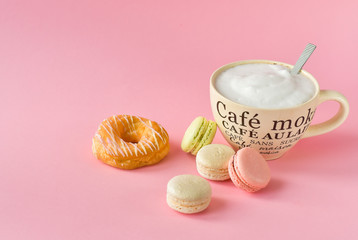 a large Cup of cappuccino coffee and donuts and a lot of colorful  French macarons, on the Cup inscription in French (mocha coffee, coffee with milk). pink background, selective focus