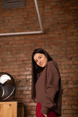 young, thin and attractive girl student dressed in casual clothes, red pants and a brown jacket, poses for a photo opposite the stone wall in the photo studio
