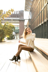 Fototapeta na wymiar Blonde junge Frau in casual Kleidung sitzt auf einer Treppe und telefoniert mit Smartphone sonnig