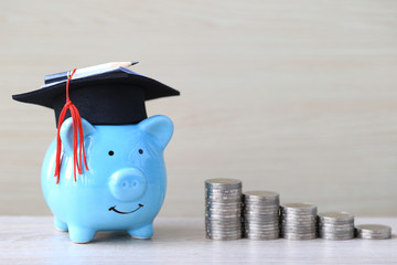 Graduation hat on blue piggy bank with stack of coins money on wooden background, Saving money for...