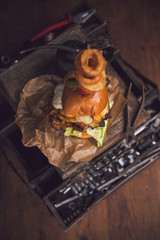 A huge stacked double cheeseburger, with bacon, cheese, lettuce, topped with gherkins, onion rings and cherry tomato. photographed in a rustic mechanic workshop background.